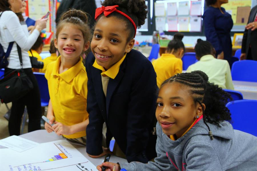 Three girls look up from a shared project to smile at the camera.